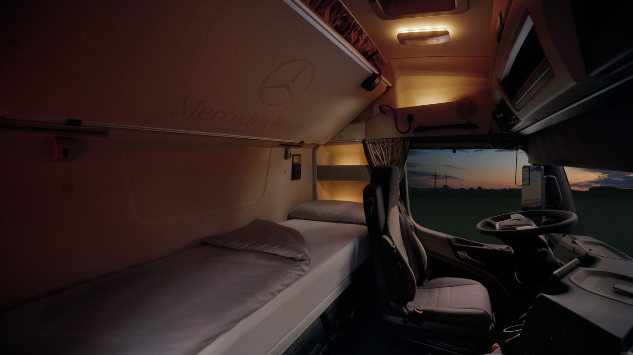 Das Bild zeigt das Innere der Kabine eines eActros von Mercedes-Benz Trucks bei Dämmerung, mit einem Blick auf das Bett und den Fahrersitz. Durch die Windschutzscheibe sind Windräder und ein Feld bei Sonnenuntergang zu sehen, was eine ruhige Atmosphäre vermittelt.