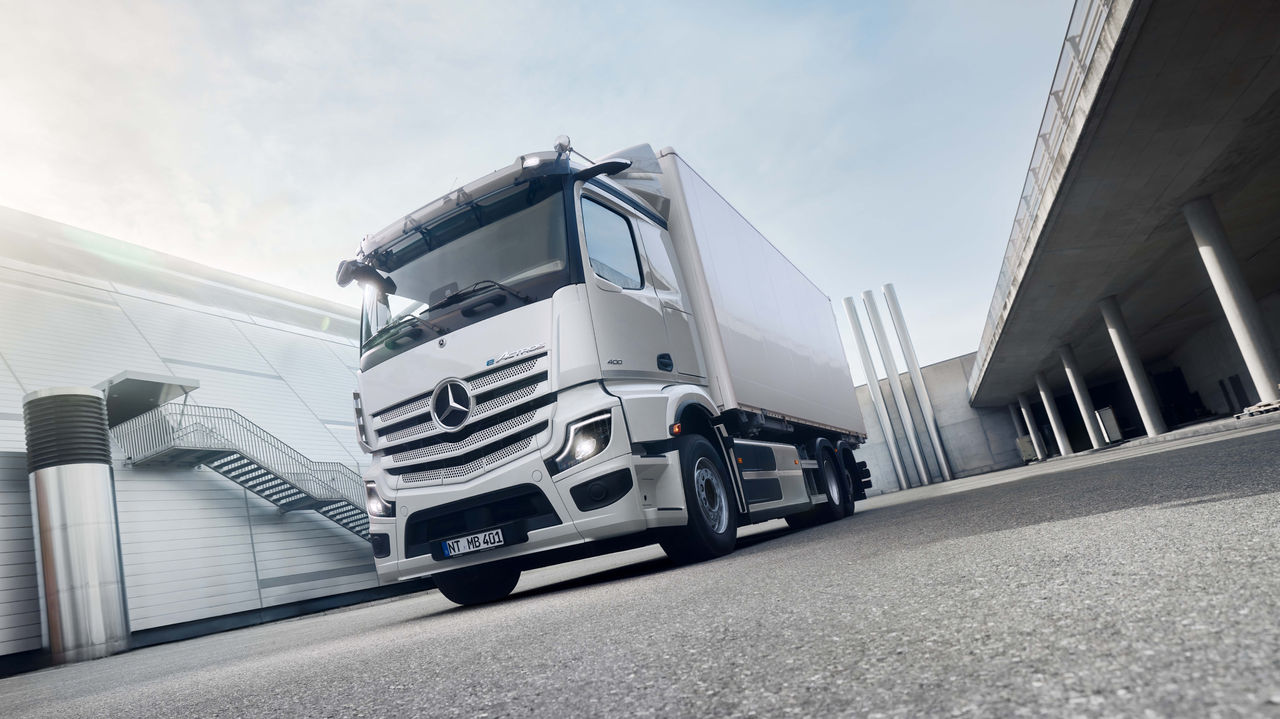 Ein weißer eActros 400 von Mercedes-Benz Trucks steht auf einer asphaltierten Fläche vor einem modernen Gebäude mit einer Außentreppe, wobei die Sonne hell von links oben scheint. Rechts im Bild ist eine erhöhte Betonkonstruktion mit Säulen zu sehen, die eine Unterführung oder einen überdachten Bereich andeutet.